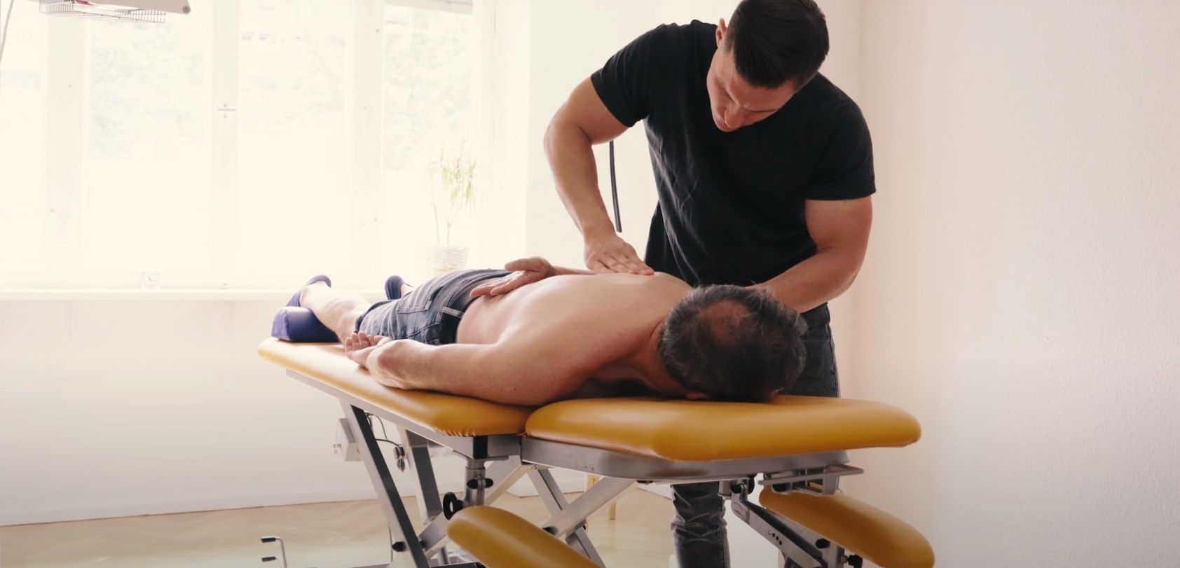 Physiotherapeutische Behandlung. Ein Mann liegt mit freiem Oberkörper auf einer Liege. Der behandelnde Therapeut ist über ihn gebeugt und löst Verspannungen im Rücken.