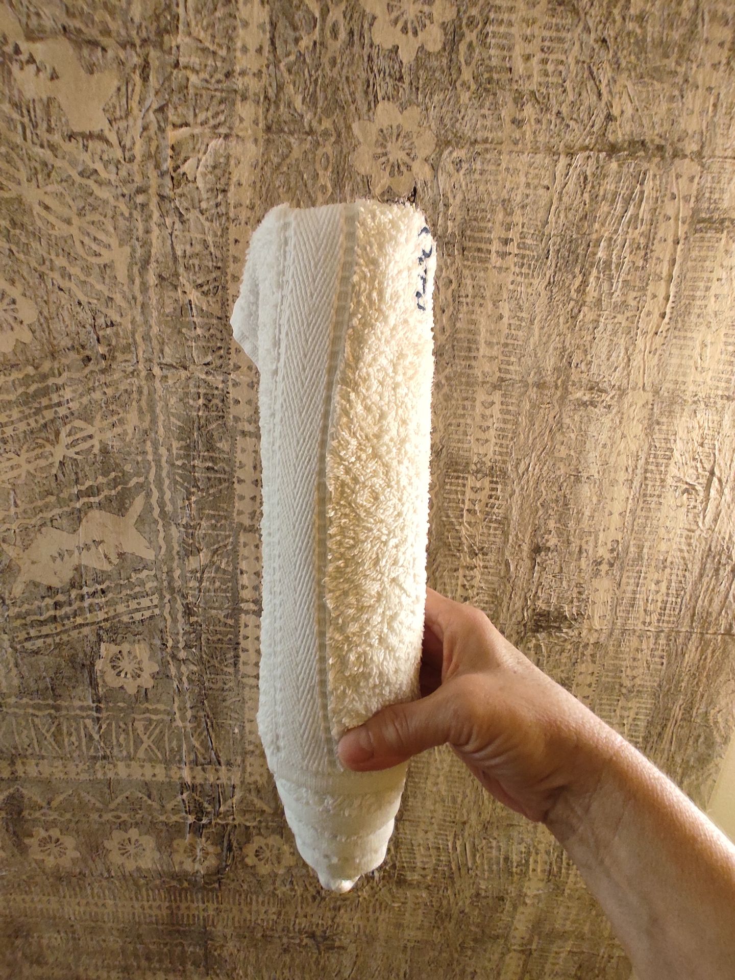 Hand hält ein zusammengerolltes weißes Handtuch vor einer gemusterten, beige-braunen Wand.