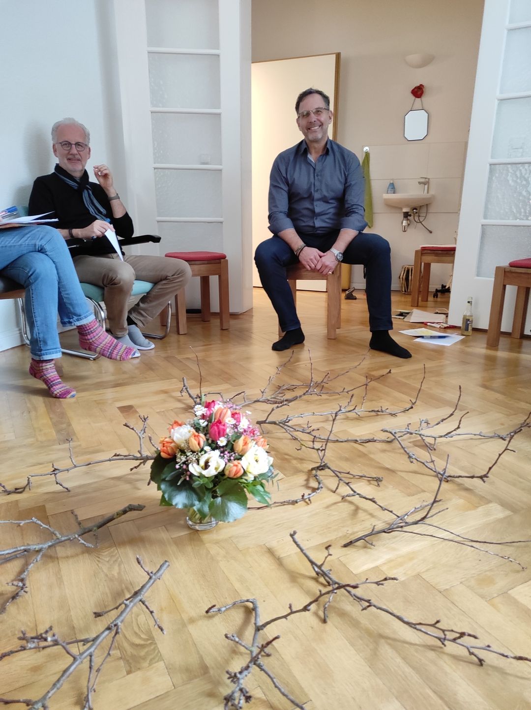 Gruppe von Menschen in einem Raum, die auf Stühlen sitzen, mit einem Blumenstrauß und Zweigen in der Mitte auf dem Boden.