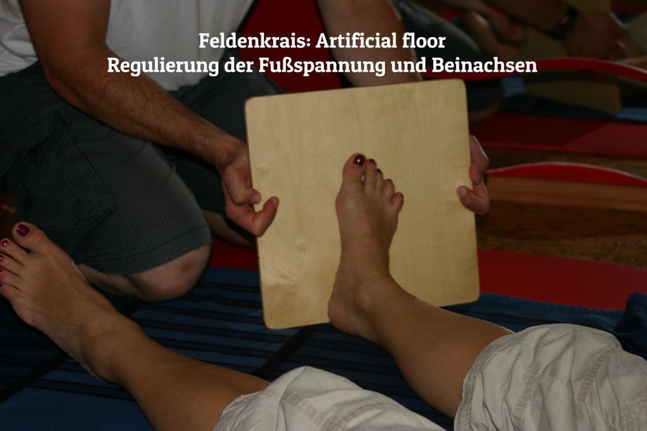 Feldenkrais-Übung zur Regulierung der Fußspannung, bei der eine Person mit dem Fuß gegen eine Holzplatte drückt, während eine andere sie hält.