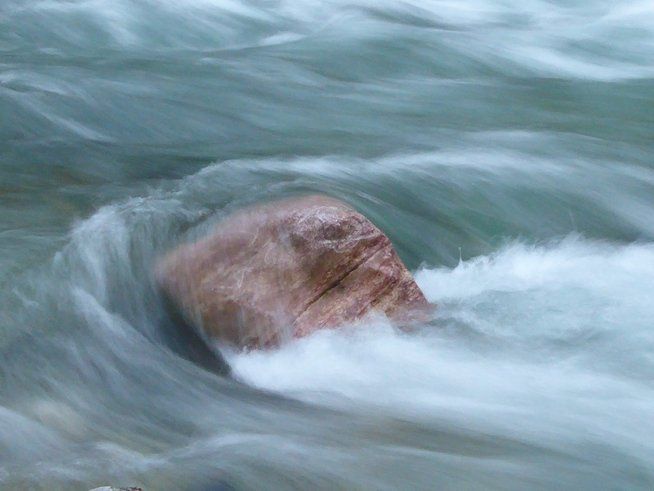 Wellen schlagen um einen braunen Felsen. Die weiße Gischt umrahmt den Stein. Das Wasser ist blau-grau und in Bewegung.