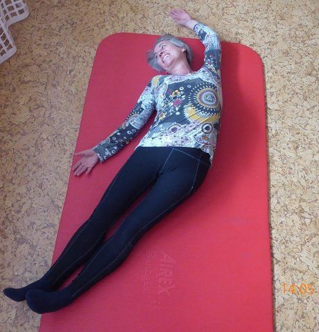 Halbmond - Ältere Frau in gemusterter Kleidung liegt lächelnd auf einer roten Gymnastikmatte und dehnt sich.