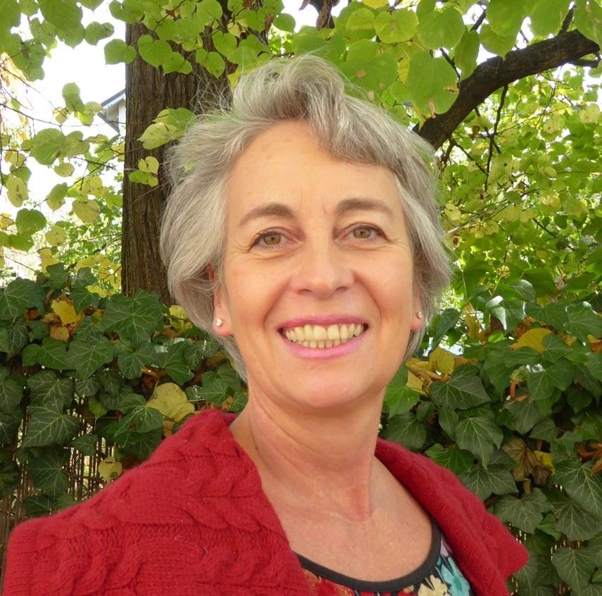 Sabrina Gußmann, mit grauem Haar und roter Strickjacke, lächelt vor einer Hecke mit grünen Blättern