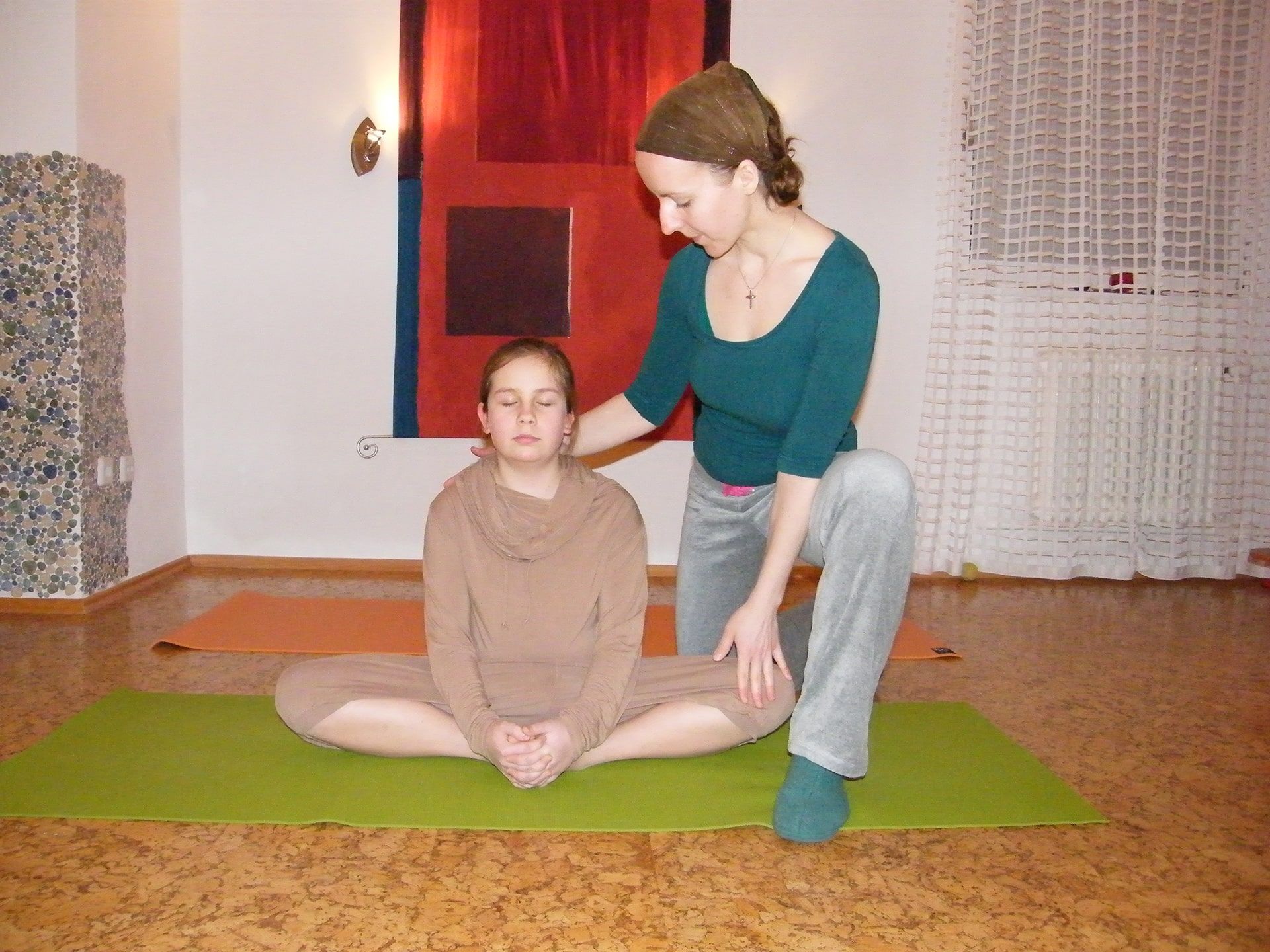 Eine Frau unterstützt ein Mädchen, die mit geschlossenen Augen im Schneidersitz auf einer grünen Matte sitzt, bei der Atemtherapie..